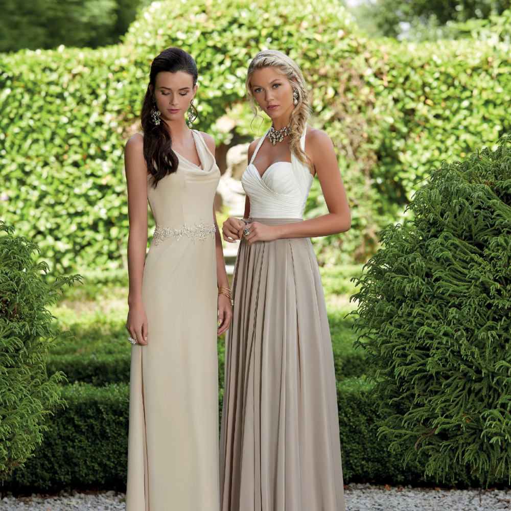 Two young bridesmaids stood by bushes in a stately home garden. One has dark brown hair and wears a long cream dress and one has blonde hair and wears a long ivory and taupe dress